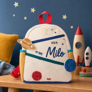 Może przedstawiać: Kremowy plecak z motywem kosmicznym, ozdobiony haftowanymi planetami, gwiazdami oraz napisem "MY HOME", "SATURN", "MARS" i imieniem "Milo" w kolorze niebieskim. Posiada czerwony uchwyt, niebiesko-żółte wykończenia oraz czerwone paski.