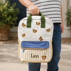 May include: A white backpack featuring embroidered brown teddy bear motifs, green top handles, and a front pocket with a blue flap. The lower section has vertical tan stripes and the name Leo embroidered in green, accompanied by a small bear icon.