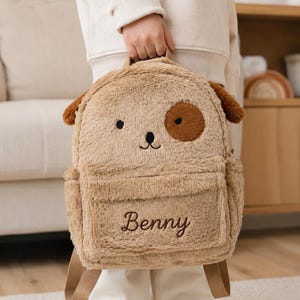 Könnte beinhalten: Ein beigefarbener Plüschrucksack in Hundeform mit Schlappohren, einem dunkelbraunen Fleck über einem Auge und gestickten Gesichtszügen. Auf der Vordertasche ist der Name Benny in dunkelbrauner Schreibschrift eingestickt. Der Rucksack hat beigefarbene Träger.