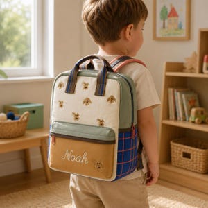 May include: A multi-colored backpack featuring an off-white upper section with embroidered dog motifs, a sage green middle trim, and a mustard yellow front pocket with the name Noah embroidered in white script. The side panel has a blue grid pattern.