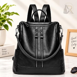 May include: A black leather-look backpack featuring two vertical zippered front pockets, a top handle, and adjustable shoulder straps. The bag has a structured design with side pockets and silver-toned metal hardware, providing a clean and functional aesthetic for daily use.