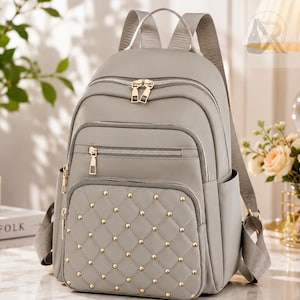 May include: A light gray backpack featuring a quilted front pocket adorned with small gold-tone metal studs. The bag includes multiple zippered compartments with gold-tone hardware, adjustable shoulder straps, and side pockets for storage.