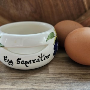 May include: A white ceramic egg separator with a hand-painted grape and leaf motif. The front of the bowl features the text Egg Separator in black script. Two brown chicken eggs are positioned next to the kitchen tool on a wooden surface.