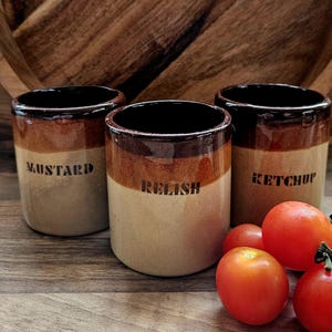 Può includere: Tre barattoli per condimenti in ceramica marrone e beige con coperchi marroni. I barattoli sono etichettati "Mostarda", "Relish" e "Ketchup". Ci sono quattro pomodori rossi davanti ai barattoli.