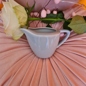May include: A light grey ceramic creamer with a handle, sitting on a pink, textured surface.