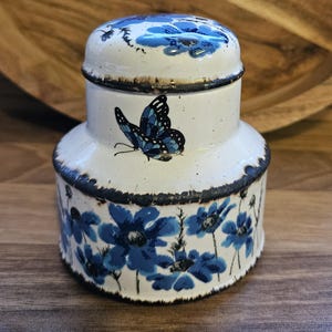 Op de afbeelding: Een witte keramische pot met een deksel, versierd met blauwe bloemen en een vlinder. De pot heeft een bruine rand en een gespikkelde witte glazuur.