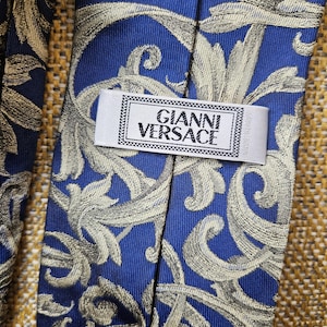 Może przedstawiać: Niebieski jedwabny krawat ze złotym kwiatowym wzorem. Krawat posiada etykietę "GIANNI VERSACE". Tło to teksturowana, złoto-brązowa tkanina. Wzór krawata jest klasyczny i elegancki.