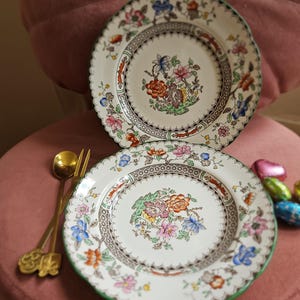 May include: Two white ceramic plates with floral patterns and green edges. The plates have a central floral design with orange, pink, blue, and green flowers. Gold-coloured fork and spoon are on the left. The plates are on a pink surface.