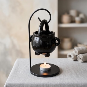 May include: A black ceramic cauldron-shaped wax warmer suspended from a curved metal stand. The glossy black pot features a handle and small feet, hanging above a circular base that holds a single lit white tea light candle for heating.