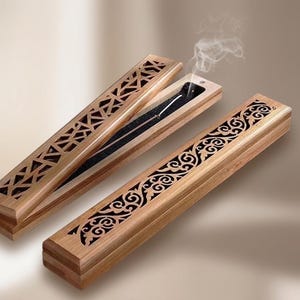May include: Two rectangular wooden incense burner boxes with intricate laser-cut patterns. One box is closed, while the other is open, revealing a black interior lining and a thin incense stick emitting a trail of white smoke into the air.