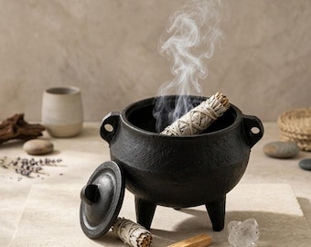 Puede incluir: Un caldero de hierro fundido negro con tres patas y dos asas laterales, con una tapa a juego apoyada al lado. Un atado de salvia blanca encendido está dentro del caldero, emitiendo un hilo de humo, junto a otro atado de salvia y un palo de madera.