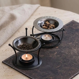 May include: Two black metal oil burners with circular frames and tripod bases. Each burner features a shallow bowl at the top for holding dried herbs or wax, and a small circular platform at the base to hold a lit tea light candle for heating.