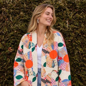 May include: A quilted jacket featuring a cream-colored base with a vibrant patchwork pattern of orange circles, green leaves, and various floral prints. The jacket has an open front, long sleeves, and a straight hem, worn over a white top and blue jeans.
