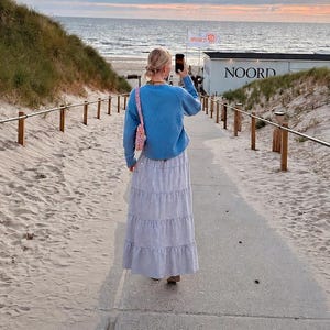 May include: A light blue long-sleeved sweater and a long, tiered, light-colored skirt. The sweater has a crew neckline and a relaxed fit, while the skirt features a subtle pattern and falls to the ankles. A small pink patterned bag is worn.