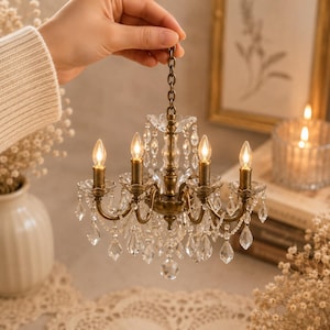 May include: A miniature chandelier featuring a metallic bronze finish, four upright candle-style light bulbs, and numerous clear hanging crystal-like teardrop prisms. The fixture is suspended by a matching bronze-toned metal chain link.