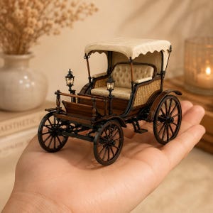 May include: A miniature model of a horse-drawn carriage featuring a dark brown wooden frame, black spoked wheels, a cream-colored tufted seat, and a matching cream fabric canopy. Two small black lanterns are mounted on the front of the carriage.