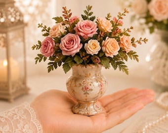 Peut inclure: Un vase miniature en céramique avec une base de couleur crème, orné de bordures dorées délicates et de motifs floraux peints. Le vase contient un arrangement dense de roses artificielles roses et pêche, entremêlées de petites feuilles vertes.