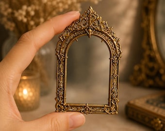 Könnte beinhalten: Ein kleiner, verzierter, goldfarbener Metallrahmen im Antik-Stil mit einem Bogen oben. Der Rahmen weist komplizierte, erhabene Filigran-Details auf, darunter florale Motive und Schnörkel entlang der Seiten sowie einen spitzen, dekorativen Abschluss.