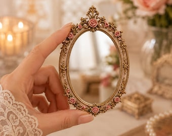 Könnte beinhalten: Ein ovaler Spiegel mit einem verzierten goldfarbenen Rahmen, der mit kleinen, geformten rosa Rosen und detaillierten Blattverzierungen geschmückt ist. Der Rahmen hat eine spitze Oberseite und eine glatte, reflektierende Glasfläche.