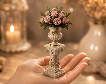 May include: A miniature decorative urn filled with a bouquet of dusty pink roses and green foliage, resting atop a classical-style square pedestal with gold-toned leaf accents. The entire structure is crafted in a light beige, marble-like finish.