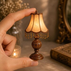 Peut inclure: Une lampe de table miniature avec une base couleur bronze aux détails sculptés complexes et un abat-jour festonné dans une teinte beige douce. L'abat-jour est orné de motifs floraux délicats et d'une frange de petites perles ambrées.