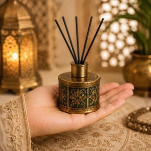 May include: A cylindrical reed diffuser with a metallic gold finish and dark green panels featuring embossed gold patterns. The diffuser has a gold-toned neck and five thin black reeds extending upward from the center of the container.