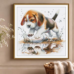 May include: A square cross-stitch embroidery pattern featuring a brown, black, and white beagle puppy jumping into a puddle. The design shows the puppy mid-air with water splashing around its paws and a clear reflection visible in the water below.