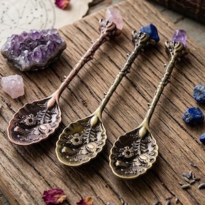 Puede incluir: Tres cucharas de metal con mangos florales ornamentados y remates de cristal en cuarzo rosa, lapislázuli y amatista. Las cucharas presentan cuencos texturizados con detalles botánicos en relieve, dispuestas junto a cristales sueltos y pétalos.
