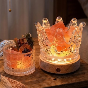 May include: A clear glass crown-shaped lamp with a light-up base containing pink salt crystals, positioned next to a matching clear glass jar filled with pink salt crystals, dried orange slices, and small pine sprigs.