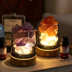 May include: Two cylindrical glass containers on light wood bases, each filled with raw crystal stones. The left container holds purple amethyst stones, while the right holds orange stones. Both are illuminated from below, accompanied by two small bottles labeled Rose.
