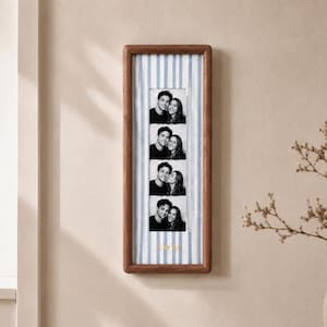Può includere: Una cornice verticale in legno contenente una striscia di quattro fotografie in bianco e nero di una coppia. Lo sfondo all'interno della cornice presenta strisce verticali azzurre e bianche, con le lettere dorate A e M separate da un cuore.