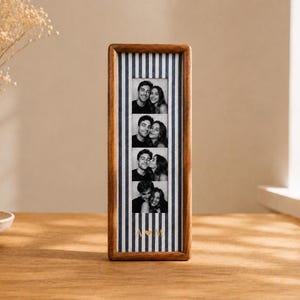 Puede incluir: Un marco de fotos rectangular de madera con un fondo de rayas verticales azules y blancas. El marco contiene una tira vertical de cuatro fotografías en blanco y negro de una pareja, con las letras doradas A y M separadas por un corazón.