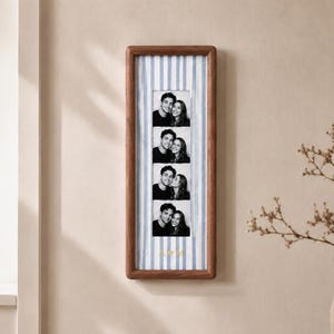 Può includere: Una cornice verticale in legno contenente una striscia di quattro fotografie in bianco e nero di una coppia. Lo sfondo all'interno della cornice presenta strisce verticali azzurre e bianche, con le lettere dorate A e M separate da un cuore.