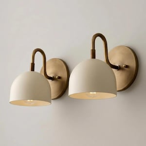 May include: Two wall sconces featuring matte cream-colored dome shades and curved brass arms. Each fixture includes a circular brass wall plate, a downward-facing semi-spherical shade, and a single light bulb socket centered within the dome.
