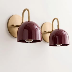 May include: Two wall sconces featuring a deep burgundy, dome-shaped shade with a glossy finish. Each lamp is attached to a circular, brushed brass wall plate by a curved, matching brass arm that holds a single light bulb inside the shade.