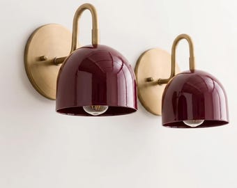 May include: Two wall sconces featuring deep burgundy dome-shaped shades and brushed brass hardware. Each fixture has a circular wall plate, a curved arm extending outward, and a single light bulb visible inside the glossy dark red metal shade.