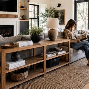 May include: A long, rectangular wooden console table with two open shelves, positioned behind a gray sofa. The table holds a beige table lamp, a black ceramic vase with greenery, several stacked books, a wooden bowl, and a woven basket.