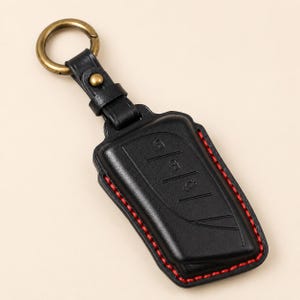 May include: A black leather car key fob cover featuring embossed button icons, red contrast stitching along the edges, and a brass-toned metal ring attached by a matching leather strap with a small brass stud fastener.