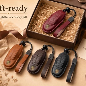 May include: Four car key fob covers in burgundy, tan, dark brown, and black, each featuring a matching leather wrist strap with a black cord and toggle. One cover is inside a brown gift box, while the others are arranged on a flat surface.