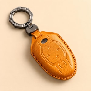 May include: An amber-colored leather car key fob cover featuring embossed button icons, including a lock, unlock, and trunk release symbol. The cover is attached to a dark gray metallic circular keyring with a mechanical, industrial-style design.