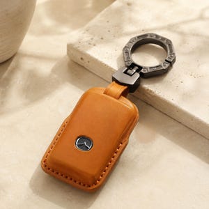 May include: A tan leather key fob cover featuring a centered silver Mazda logo emblem. The rectangular case is stitched along the edges and connects to a dark metallic, circular, segmented key ring via a matching dark metal hinge mechanism.