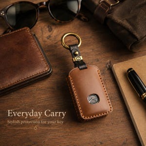 May include: A brown leather key fob cover featuring a metal Mazda logo emblem, a black leather strap, and a brass-toned circular keyring. The text "Everyday Carry" and "Stylish protection for your key" is printed in white on the lower left side.