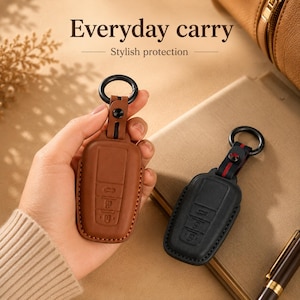 Peut inclure: Deux étuis en cuir pour clé de voiture, un marron clair et un noir, avec des icônes de boutons en relief, des bords cousus et un anneau métallique noir. Le texte Everyday carry Stylish protection est visible en haut de l'image.