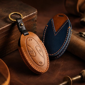 Peut inclure: Deux étuis à clés ovales en cuir avec des coutures apparentes. L'un est marron clair avec un anneau en laiton fixé par une lanière en cuir noir. L'autre est bleu foncé avec une pièce d'accent en cuir marron clair sur la partie supérieure.
