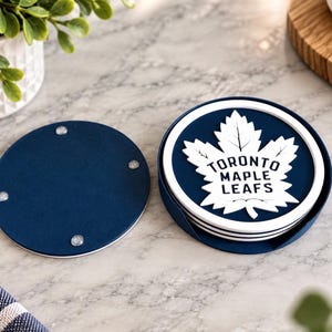 May include: A set of round drink coasters featuring the Toronto Maple Leafs logo in white on a dark blue background. The coasters are stacked inside a matching dark blue circular holder, with one coaster placed face down showing four clear feet.