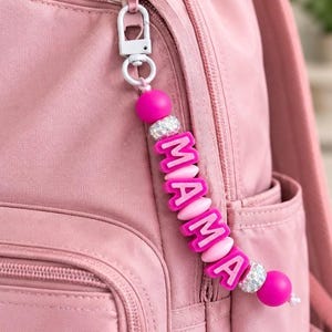 May include: A pink backpack featuring a decorative keychain attached to the zip. The keychain consists of bright pink spherical beads, two silver-toned rhinestone-encrusted spacer beads, and pink letter beads spelling out the word MAMA.