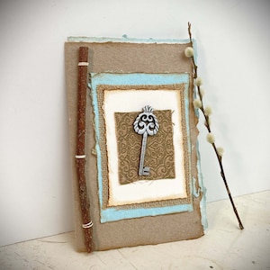 May include: A handmade card with a rustic brown paper background and a blue and white patterned frame. A silver key with a decorative design is centered in the frame.