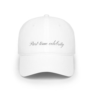 May include: A white baseball cap featuring the text Part-time celebrity printed in a black, elegant, cursive script font across the front center panel. The cap has a structured crown, a curved brim, and a button at the top center of the dome.