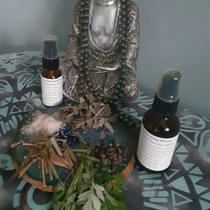 May include: A silver Buddha statue sits on a table with a small bottle of "Bewitching Brew" smudge spray, a bottle of "Witch's Bewitching" oil, and a small bowl of herbs and crystals. The text on the bottles reads "Bewitching Brew" Smudge Spray and "Witch's Bewitching" Oil.
