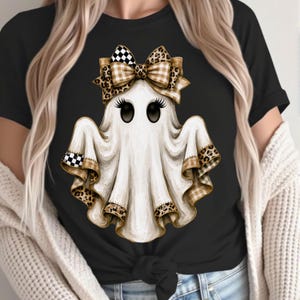 May include: A black t-shirt featuring a central graphic of a white ghost with long eyelashes. The ghost wears a large bow on its head and has ruffled edges on its base, both decorated with a mix of tan plaid, leopard print, and black-and-white checkerboard patterns.